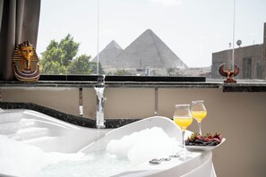 A bathtub with a view of pyramids, a decorative headpiece, and fruit on a tray. - Camel Pyramids View Inn (Giza)