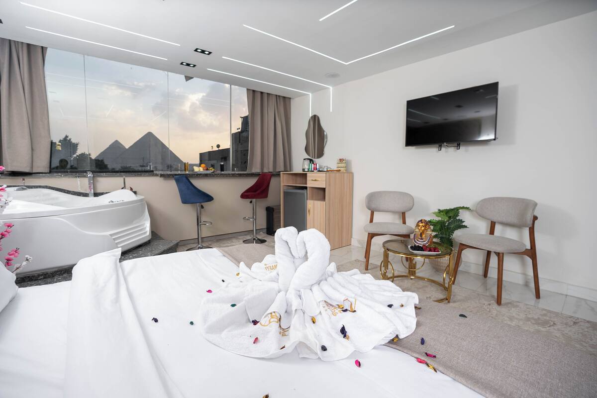 Habitación de hotel moderna con una cama grande, un jacuzzi, un escritorio con televisor y vistas a las pirámides a través de la ventana.