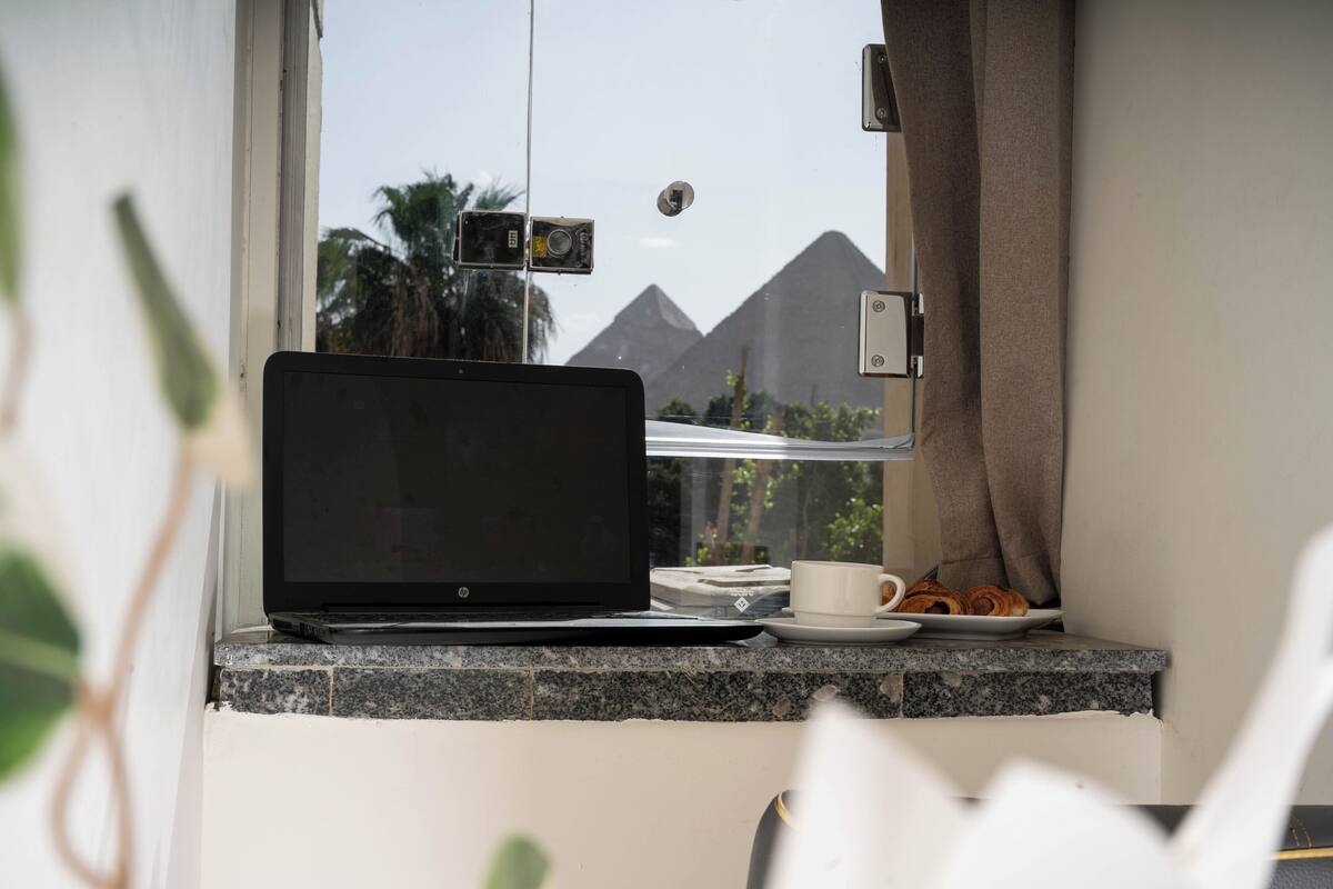 Una laptop sobre un alféizar con vista a pirámides, una taza y un plato de pasteles.