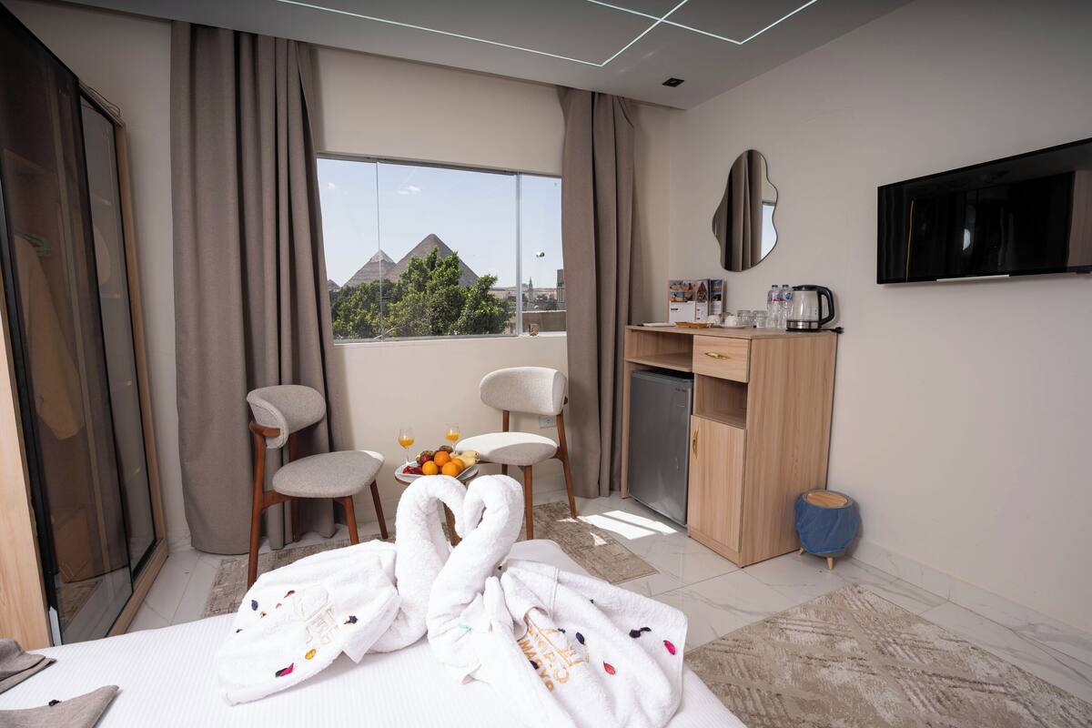 Habitación de hotel con una cama, dos sillas, un escritorio con cafetera, televisión y vistas a las pirámides.