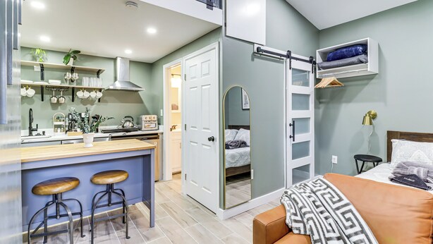 Un espace de vie compact comprenant un coin cuisine, un coin repas avec des tabourets, un lit agrémenté d’un couvre-lit à motifs et une petite table de chevet avec une lampe.