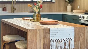 Eine Küche mit Holzausstattung, einer zentralen Kochinsel mit gewebter Tischmatte, einer Vase mit Blumen und einem Fenster mit Blick auf Bäume.