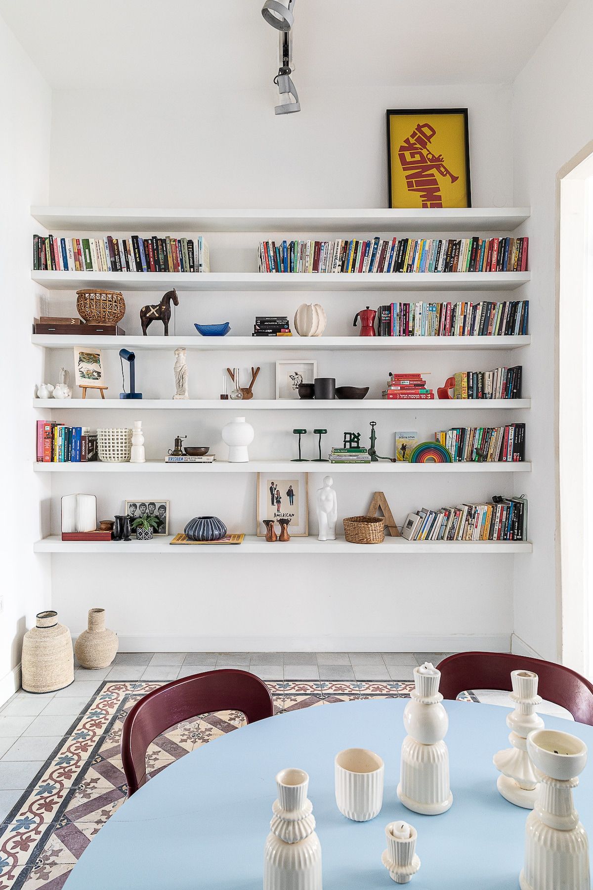 Ein Zimmer mit einer weißen Bücherwand, gefüllt mit Büchern und Dekorationsgegenständen, einem Esstisch mit weißen Keramikobjekten und einem gemusterten Teppich.