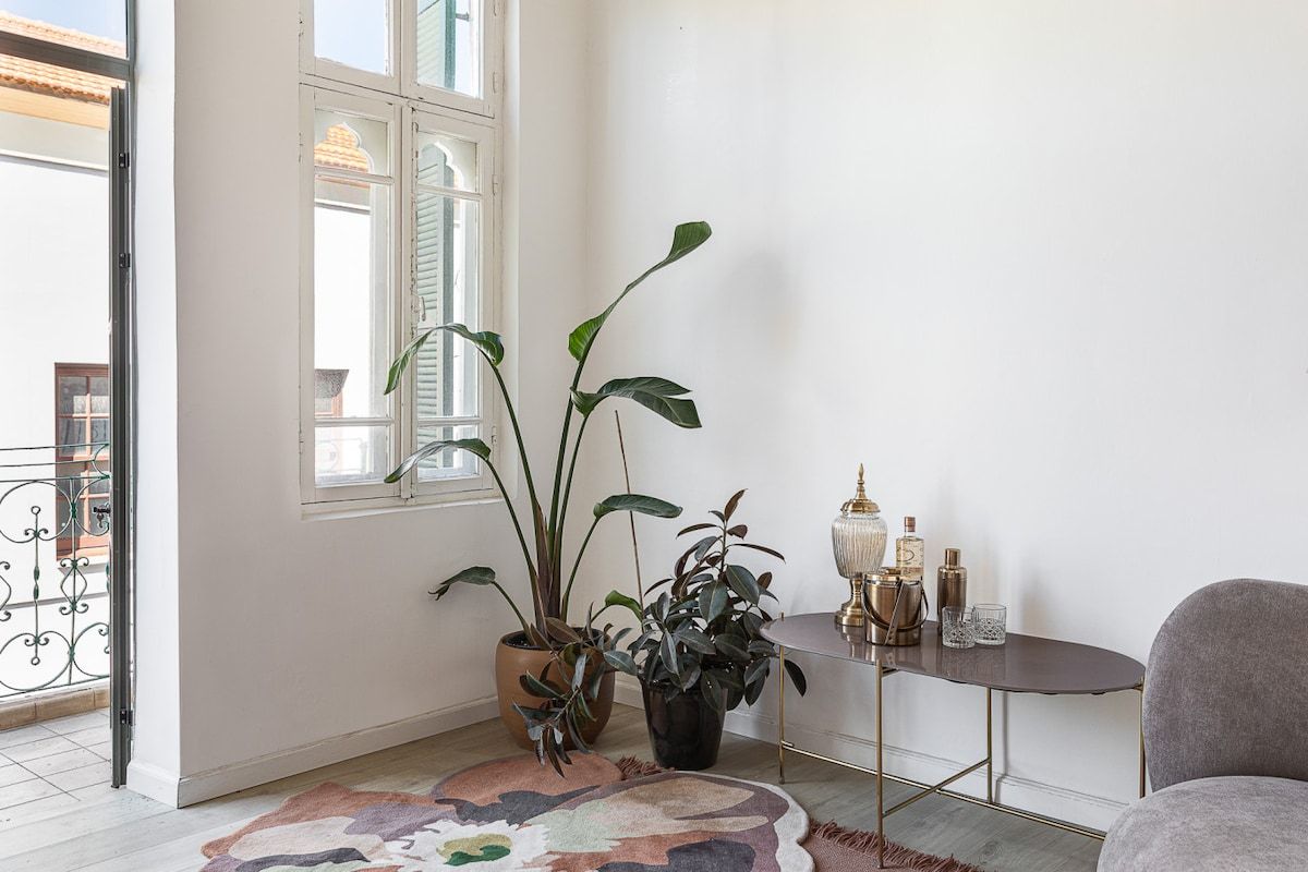 Ein Zimmer mit einem großen Fenster, einem Blumentopf, einem kleinen runden Tisch mit einer Lampe und einem Sessel.