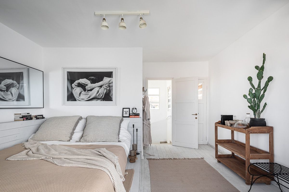 Une chambre à coucher avec un lit, deux photographies encadrées, une table de chevet, une petite table en bois, un cactus en pot et une porte donnant sur une autre pièce.