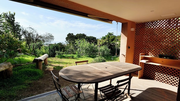 An outdoor patio with a wooden table and benches, surrounded by lush greenery and a view of the sea.