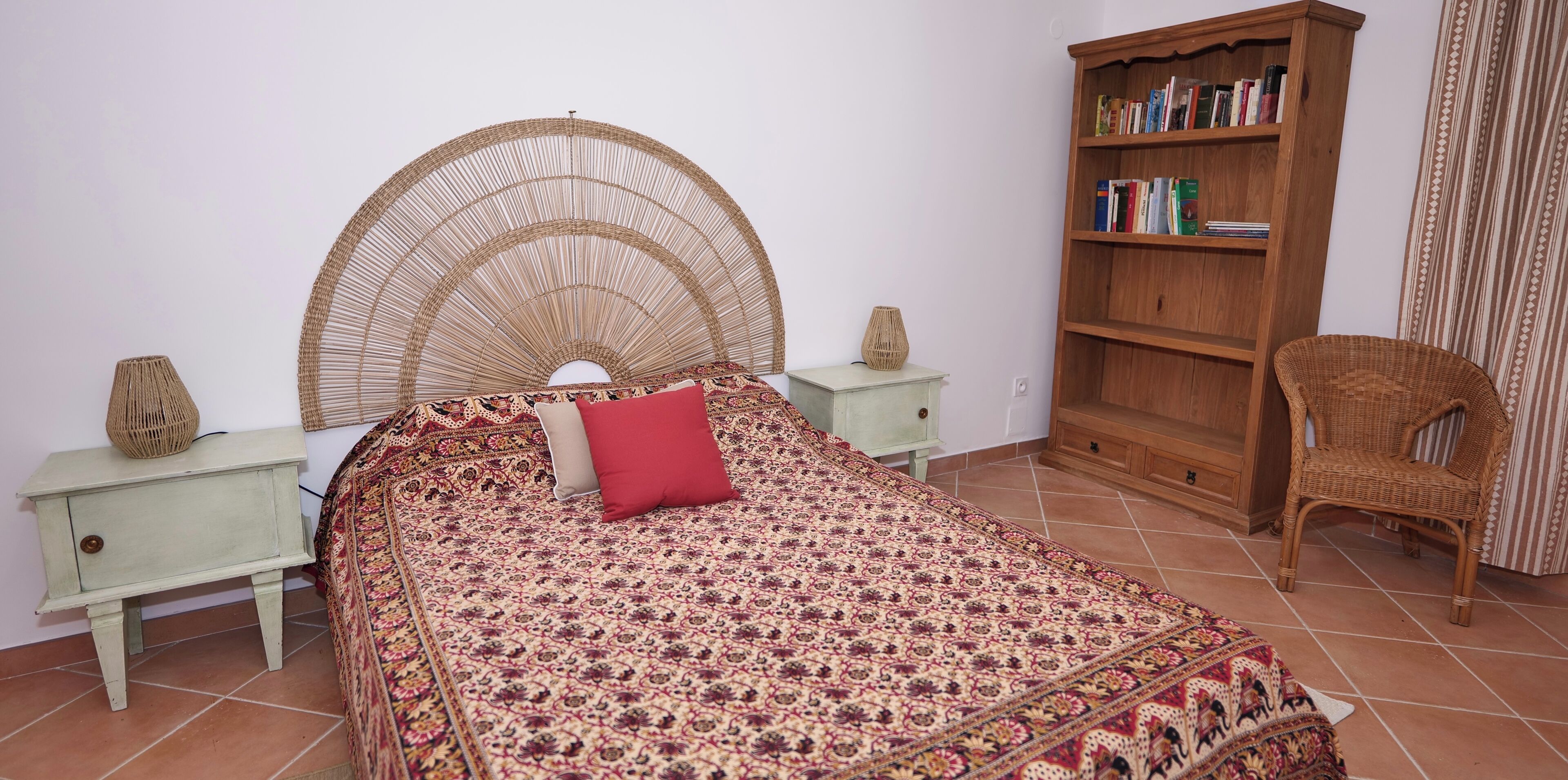 A bedroom with a bed, a wooden bookshelf, a wicker chair, and a small table with a lamp.