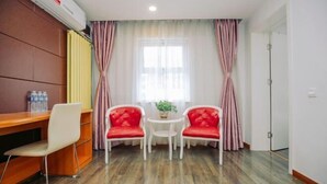 Una habitación con un escritorio de madera, dos sillas rojas, una pequeña mesa blanca con una planta y una ventana con cortinas.