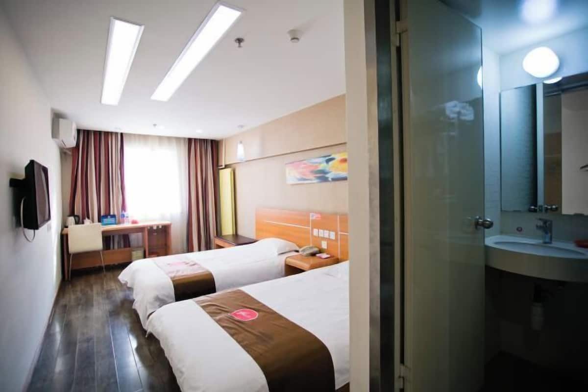 A hotel room with two beds, a desk, a chair, a TV, and a bathroom with a sink and mirror.