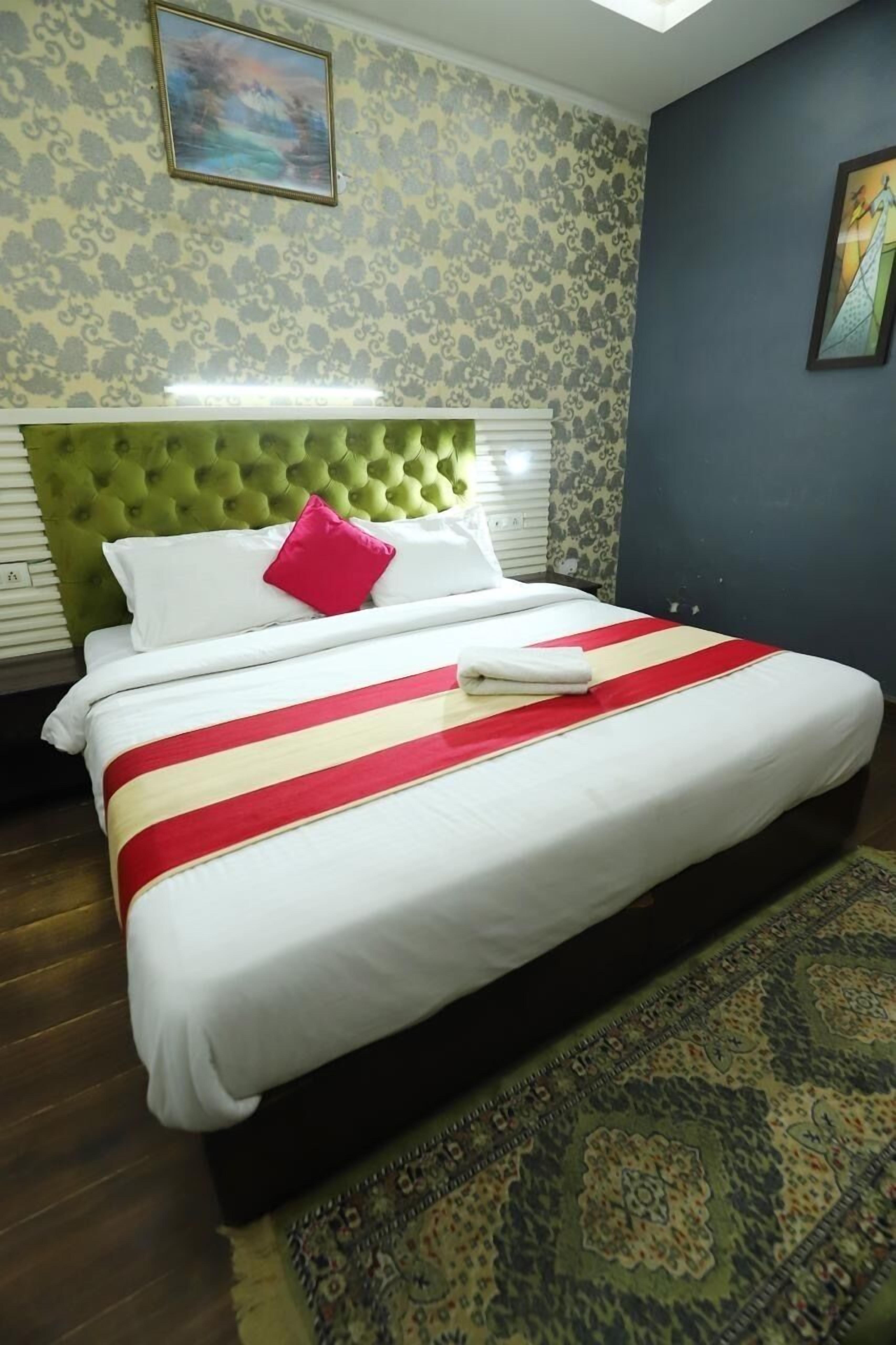 Una habitación de hotel con cabecera verde tapizada, ropa de cama blanca, colcha beige y roja a rayas, almohada roja y alfombra estampada.
