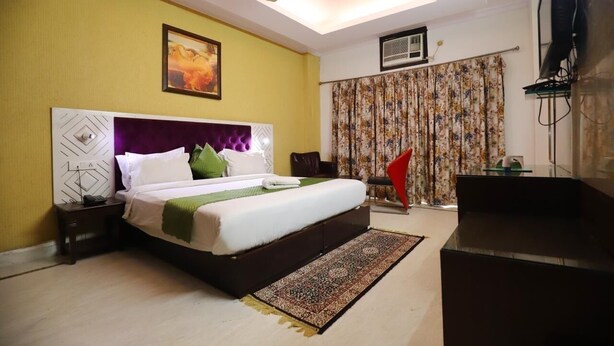 A bedroom with a bed, a nightstand, a ceiling fan, a TV, and a window with curtains. - The Hotel Vagmi (Gurugram)