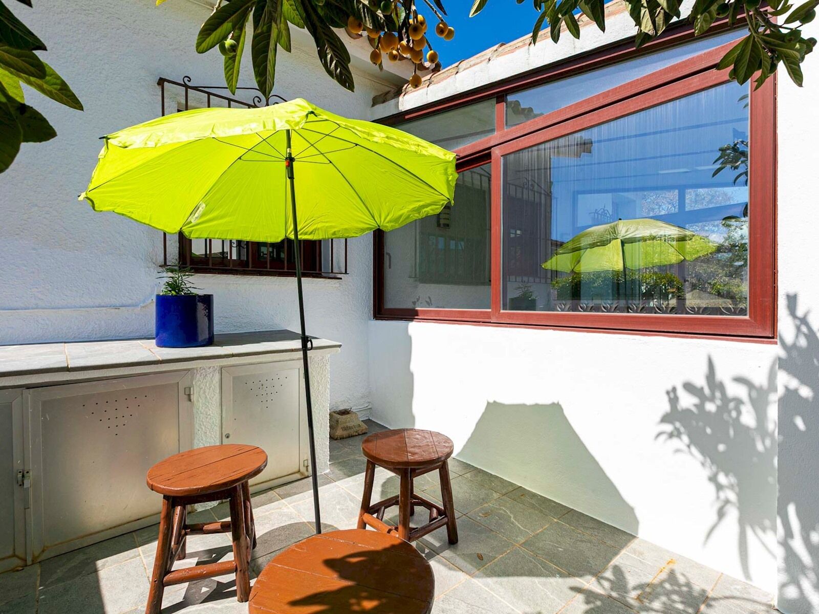 En lille patio med et bord og to skamler, en lys gul parasol og et vindue med refleksioner af andre parasoller.