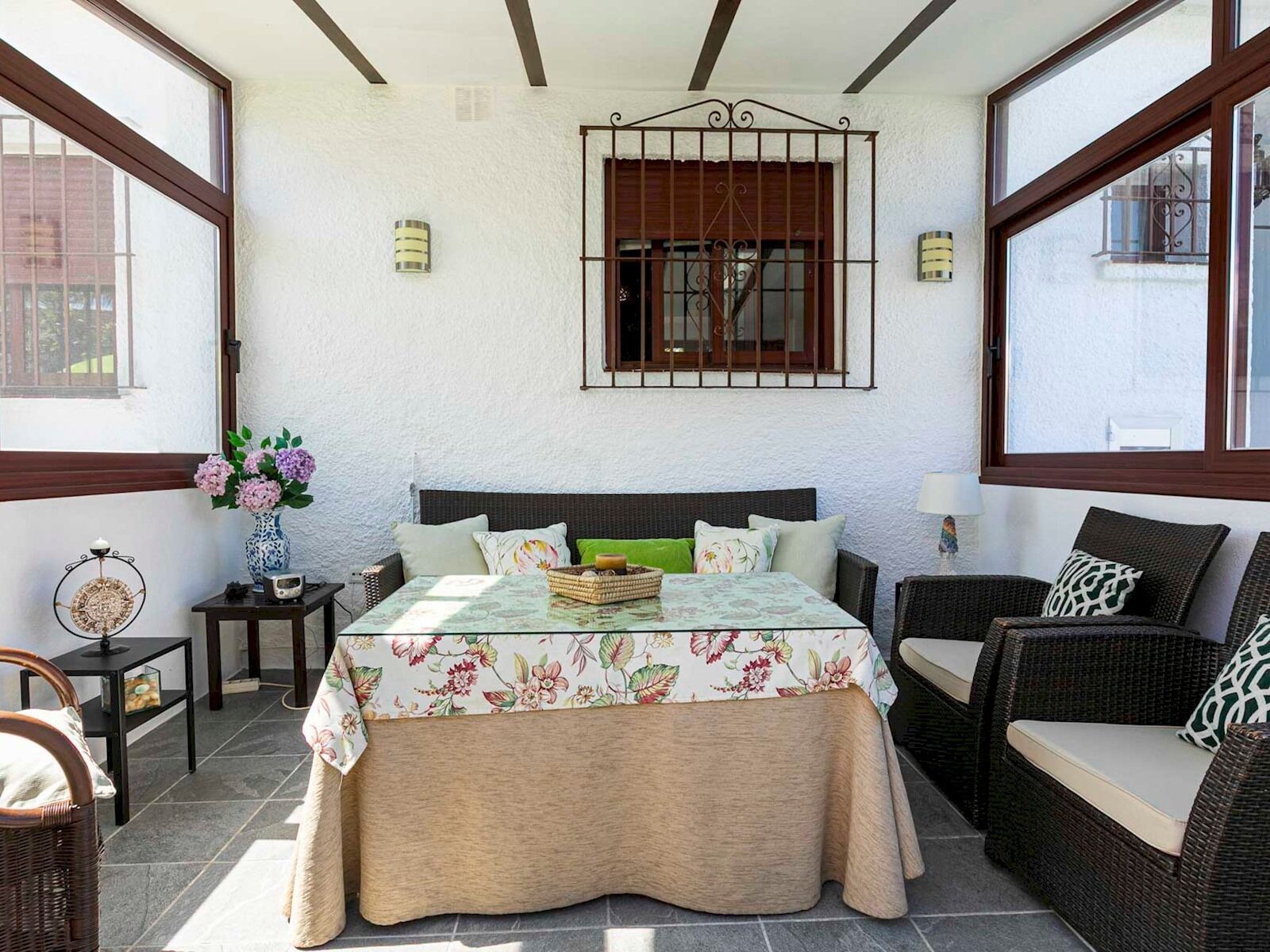 A bedroom with a bed, a small table, a wicker chair, and a window with a metal gate.