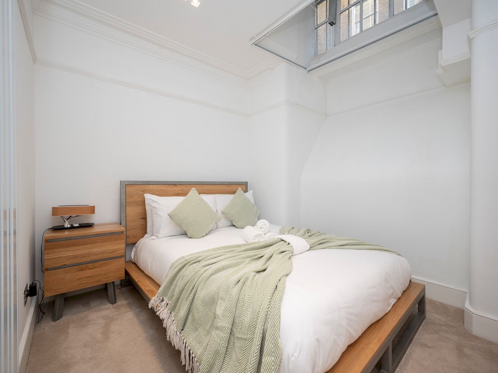 A bedroom with a wooden bed, white bedding, a bedside table with a lamp, and a window with a view.
