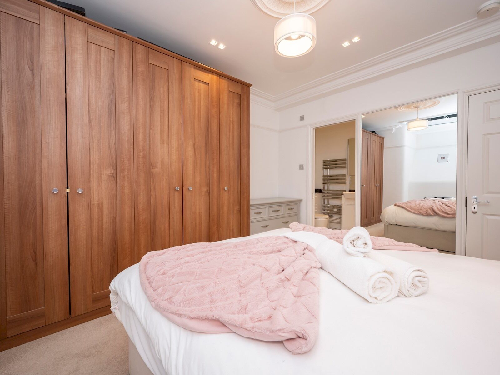 A bedroom with a large wooden wardrobe, a bed with pink bedding, and a mirror reflecting the room.
