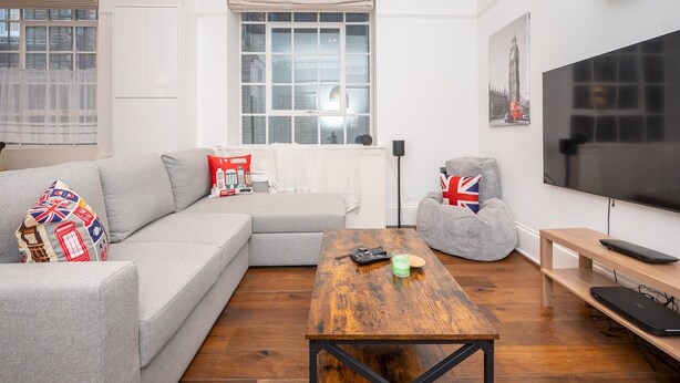 Ein modernes Wohnzimmer mit grauem Sofa, einem Holztisch, einem grauen Sitzsack, einem Flachbildfernseher und einem Fenster mit Blick auf Gebäude.