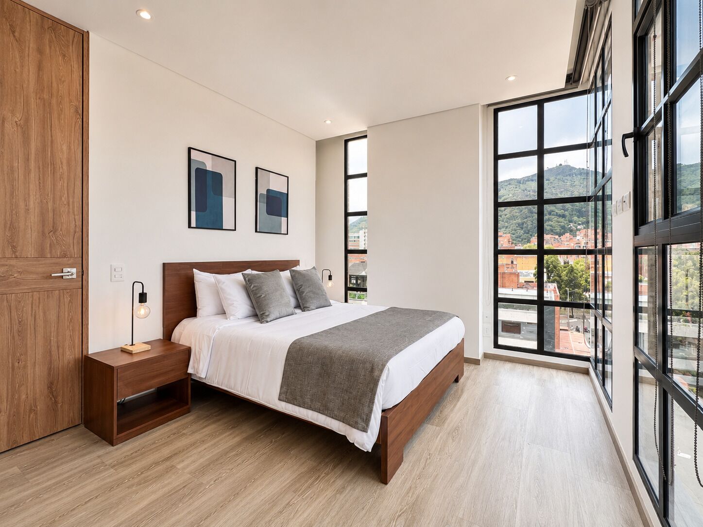 A modern bedroom with a large bed, wooden floor, and a city view through large windows.