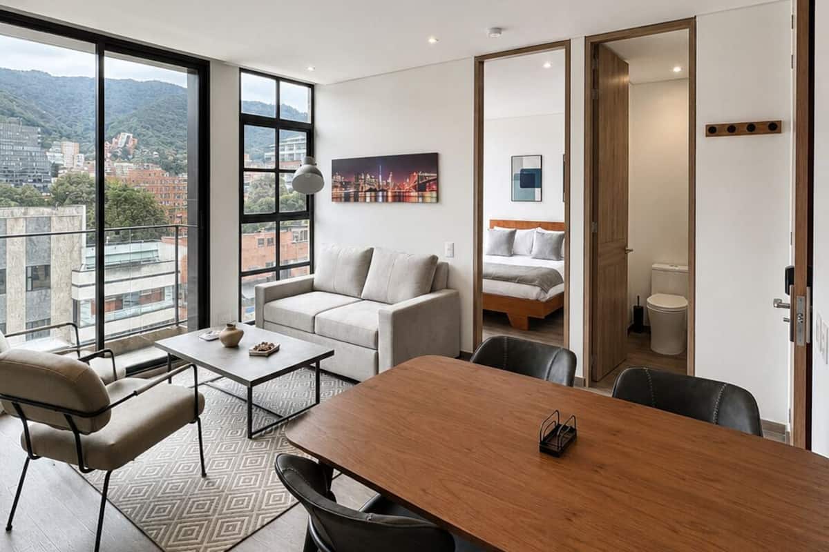 A modern living room with a large window, a sofa, a dining table, and a view of a cityscape.