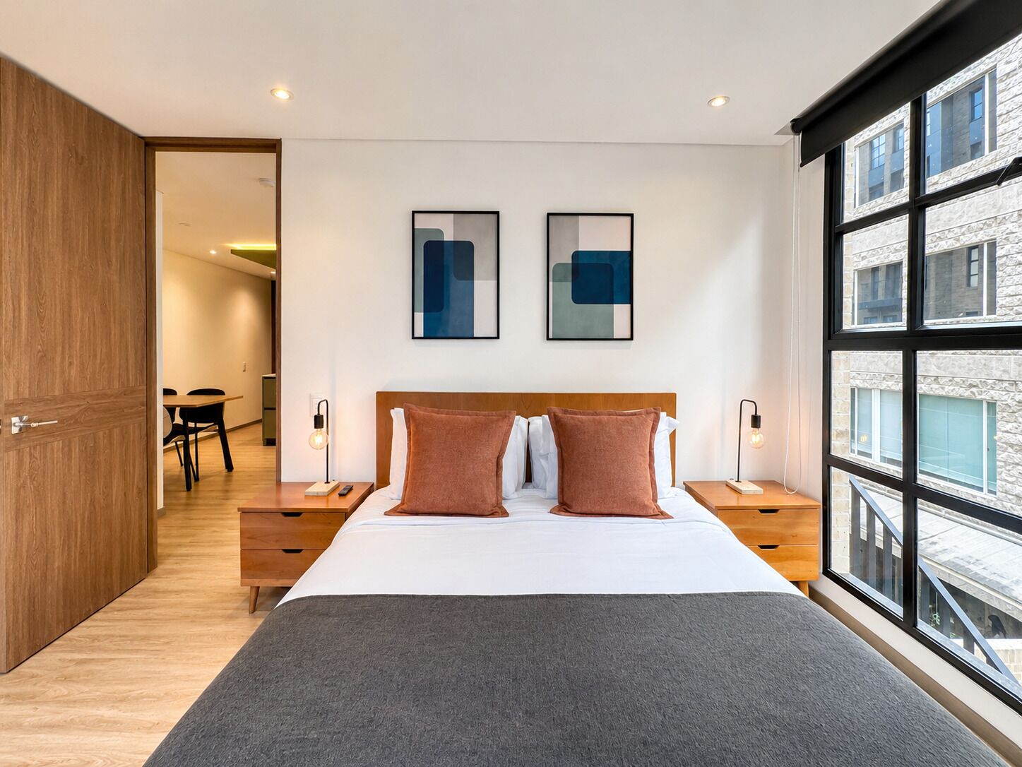 A modern hotel room with a large bed, two bedside tables, a wooden wardrobe, and a large window offering a city view.