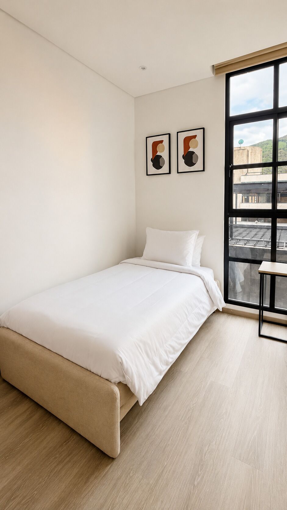 Un dormitorio minimalista con una cama individual, una ventana que ofrece vistas de edificios y dos obras de arte abstractas en la pared.