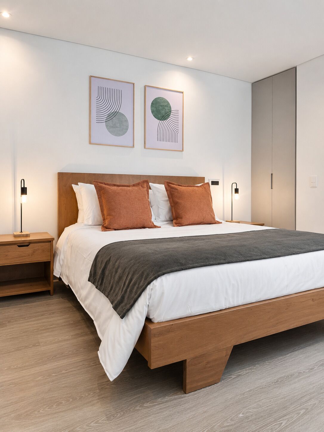 Un dormitorio moderno con cabecera de cama de madera, ropa de cama blanca, dos almohadas, una manta gris y dos obras de arte colgadas en la pared.