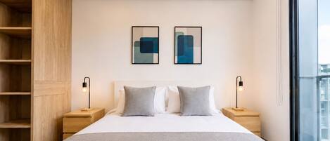 A modern bedroom with a large bed, two bedside tables, a built-in wooden shelving unit, and a large window.