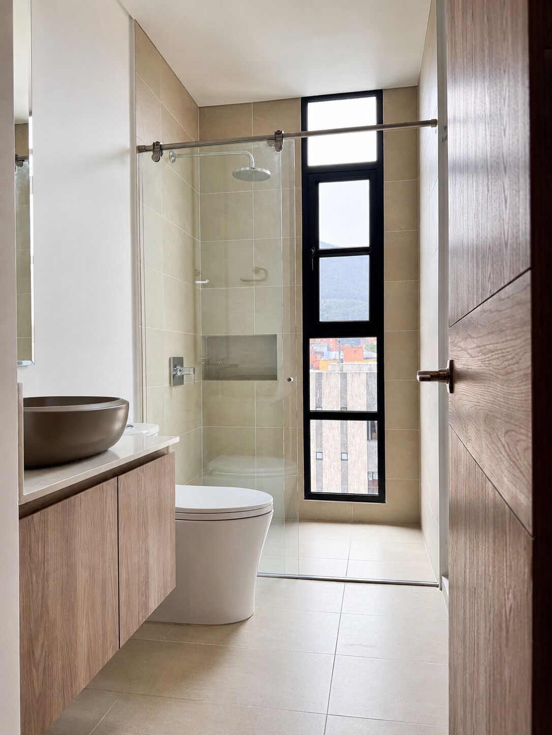 A modern bathroom with a glass shower enclosure, a large window, and a wooden vanity.