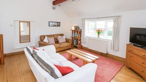 A cozy living room with a sofa, a wooden bench, a bookshelf, a TV, and a window with curtains. - The Barn - Poplar Farm (York)