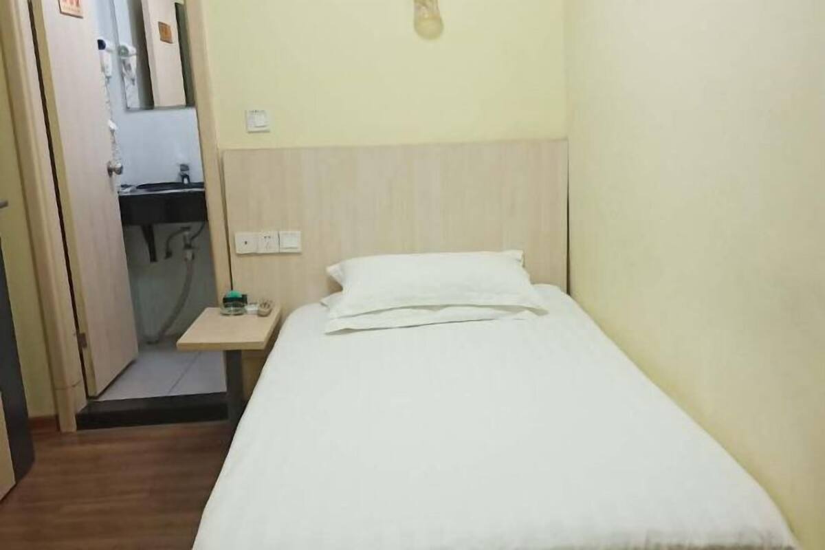 A small, single-bed room with a wooden floor, a bedside table with a lamp, and a door leading to a bathroom.