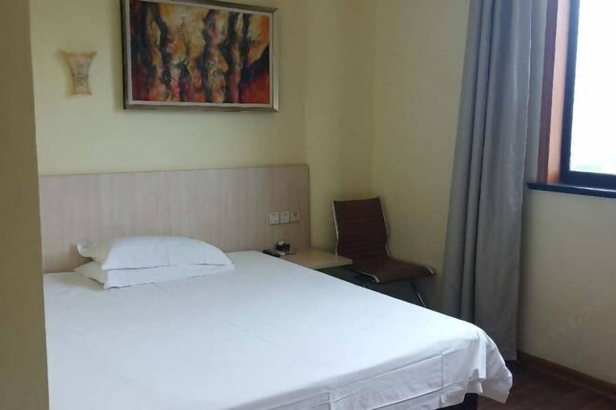 A hotel room with a bed, a chair, a painting on the wall, and a window with curtains.