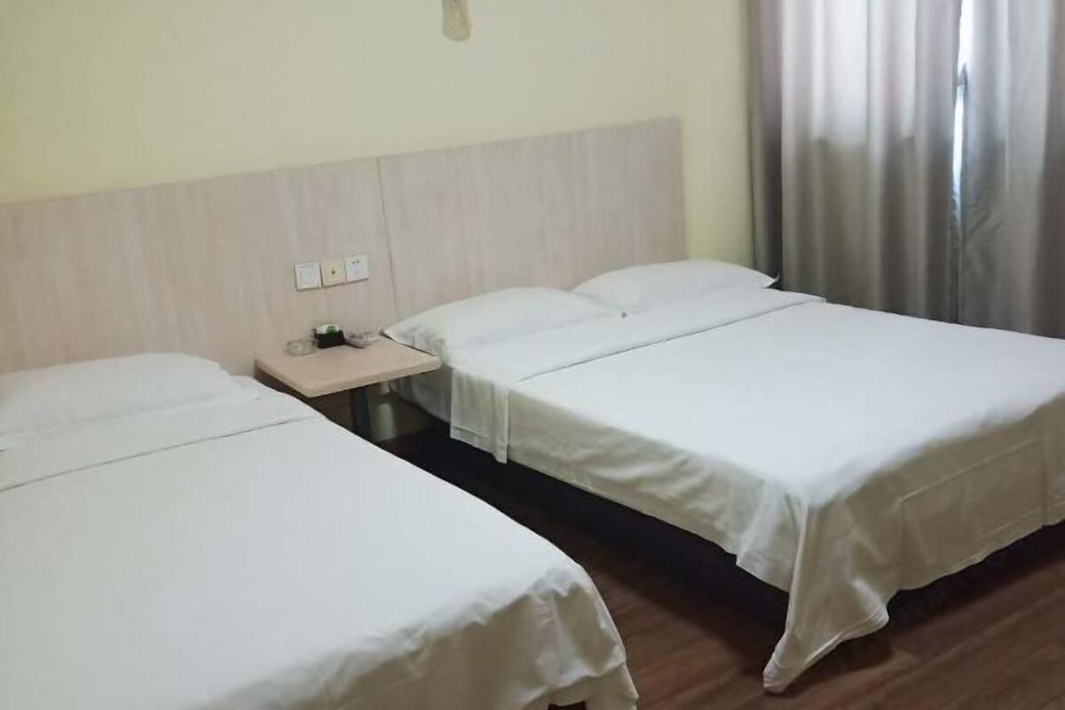 A hotel room with two single beds, a wooden headboard, a nightstand with a lamp, and a window with curtains.