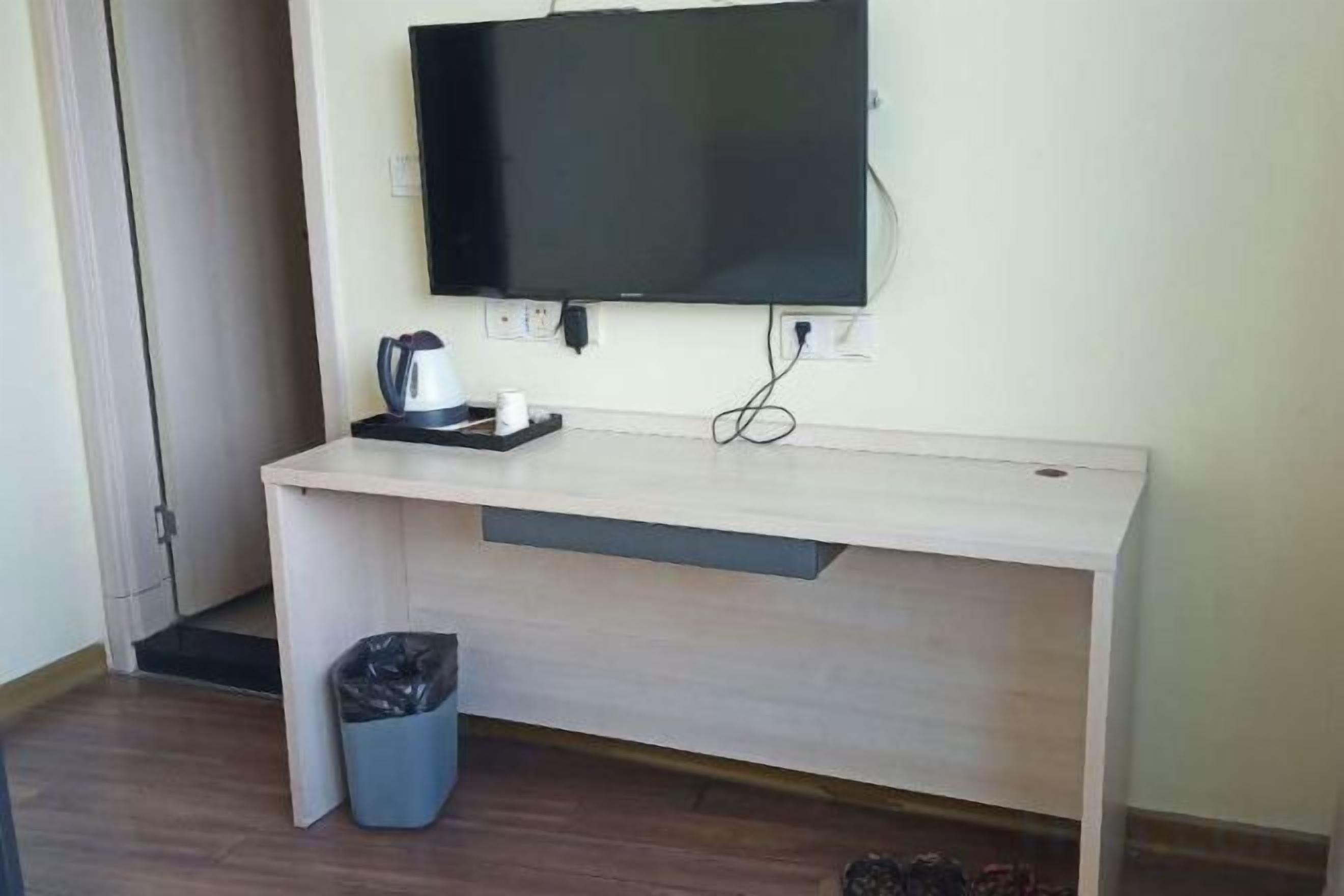 Una habitación con un escritorio de madera, un televisor montado en la pared, un cubo de basura y una cafetera.
