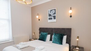 Ein ordentlich bezogenes Bett mit weißen Leinentüchern und türkisblauen Kissen, ein Nachttisch mit einer Lampe und ein gerahmtes Kunstwerk an der Wand.