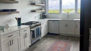 A modern kitchen with white cabinetry, a gas stove, a coffee maker, and a window with a view of greenery. - Sea Bird 3/3 Overlooking the Sea of Abaco with Private Beach & dock access (green turtle cay)