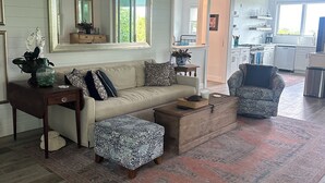 A spacious living room with a large sofa, a coffee table, and a patterned ottoman. There is a kitchen visible through an open doorway. - Sea Bird 3/3 Overlooking the Sea of Abaco with Private Beach & dock access (green turtle cay)