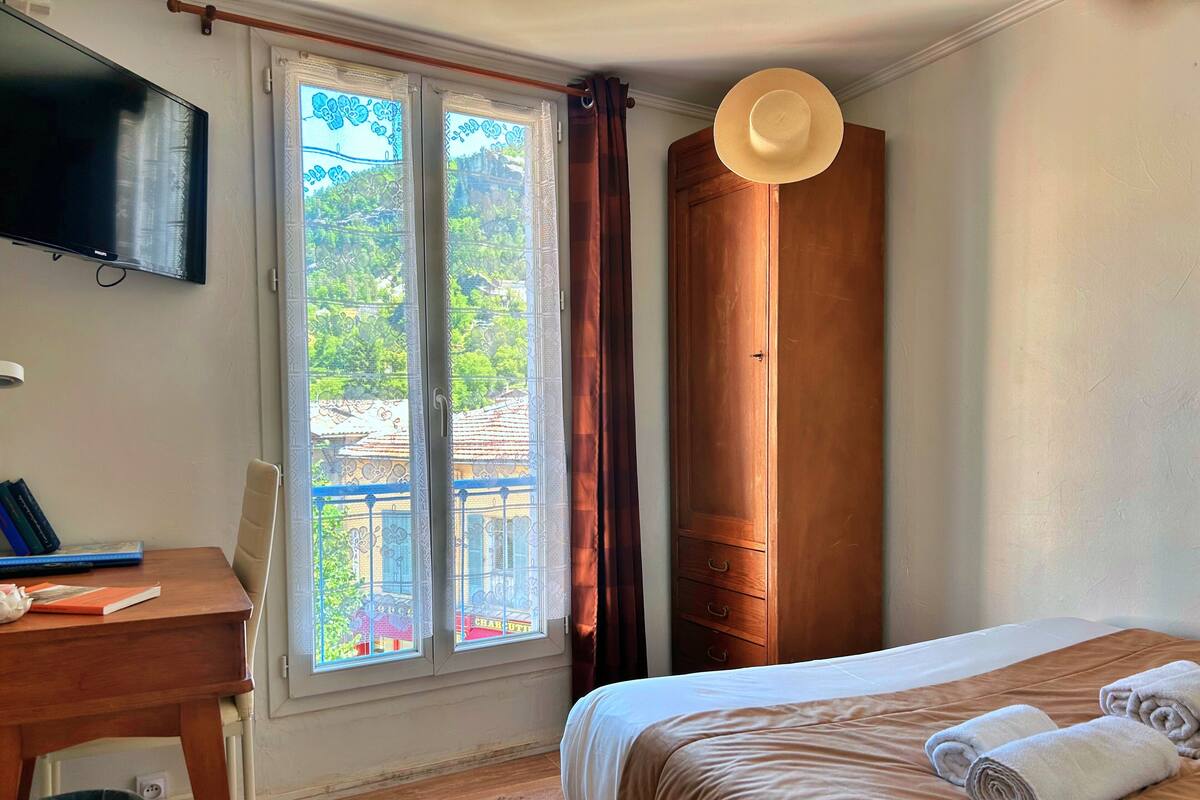 A hotel room with a bed, a desk with books, a television, and a window with a view of buildings.
