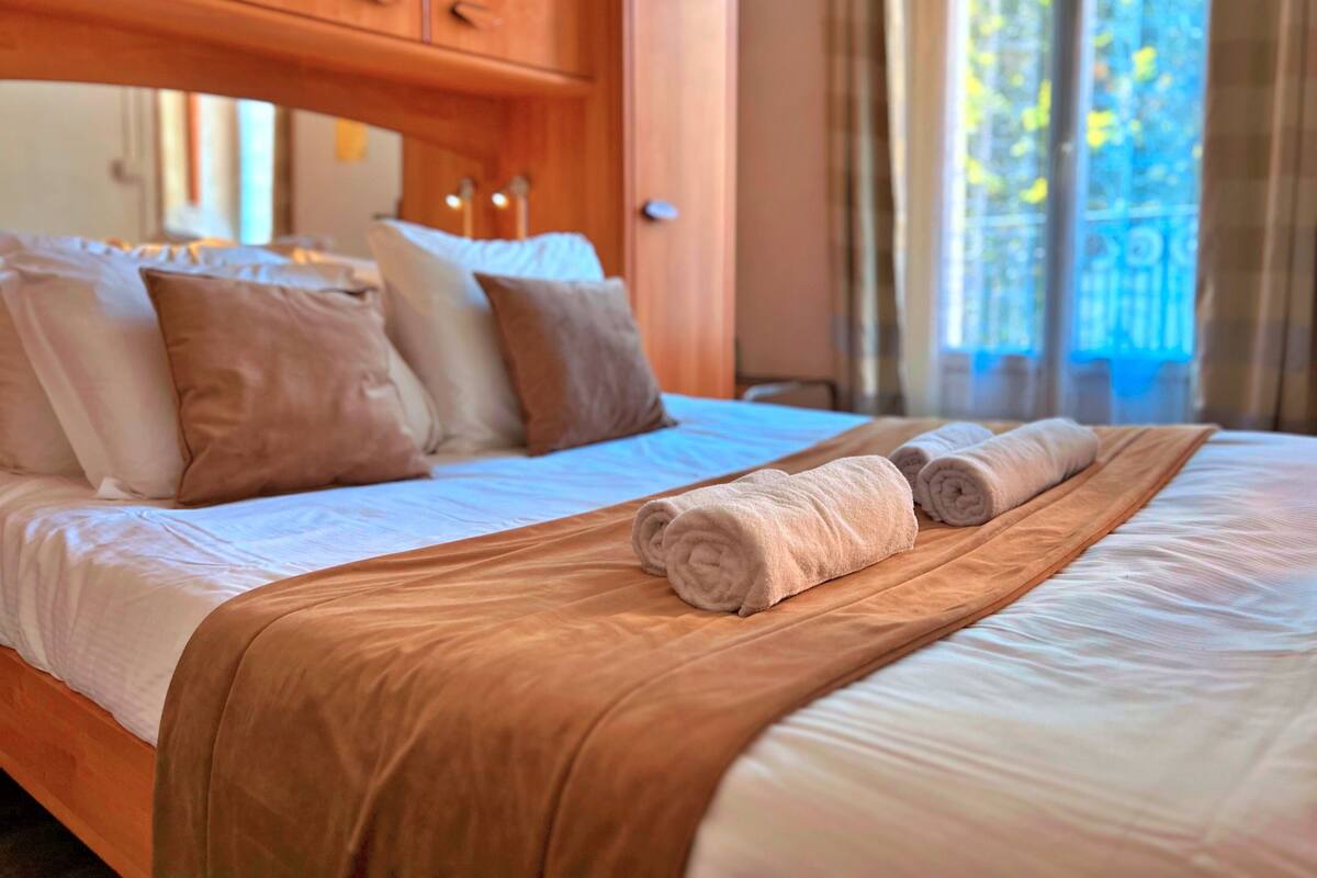 A neatly made bed with a brown bedspread and two rolled towels, wooden headboard, and a window with curtains.