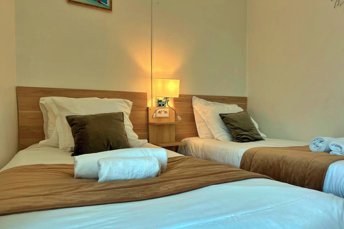 A hotel room with two beds, each with a wooden headboard, white linens, and brown pillows. A nightstand with a lamp and a power outlet is between the beds.