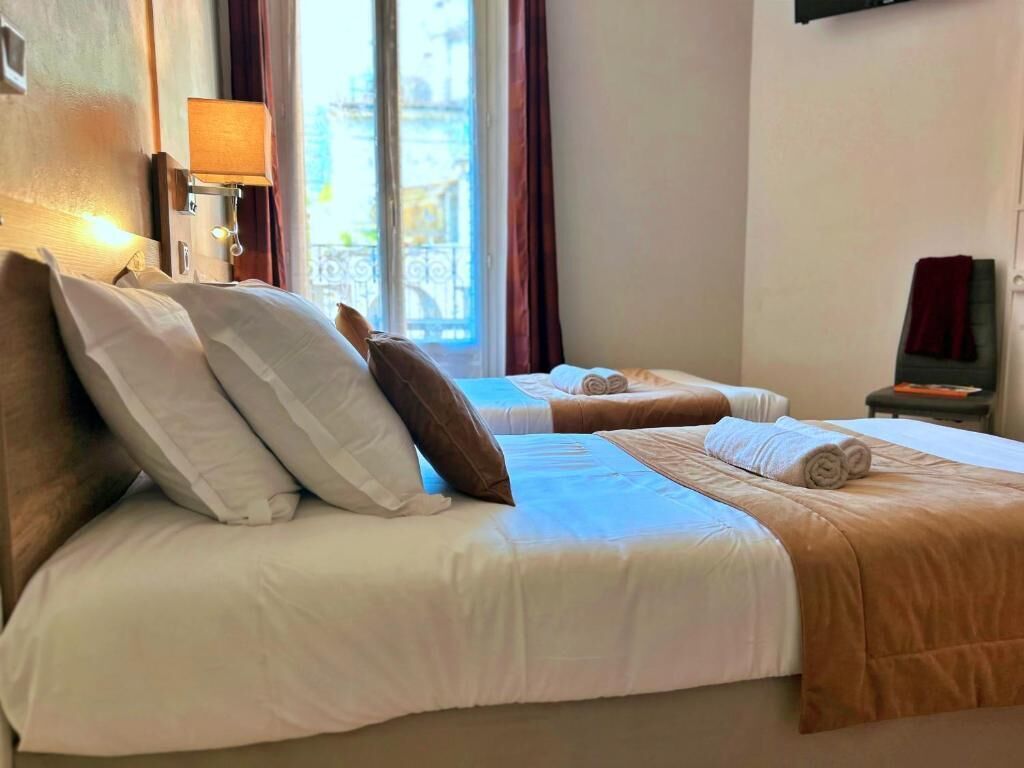 Habitación de hotel con dos camas, ventana con cortinas, lámpara y vista a la ciudad.