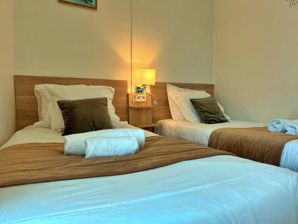 A hotel room with two beds, each with a wooden headboard, white linens, and brown throw blankets. There is a nightstand with a lamp between the beds.