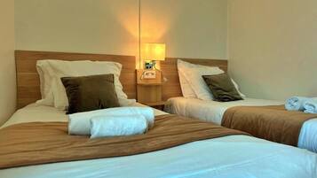 A hotel room with two beds, each with a wooden headboard, white linens, and brown throw blankets. There is a nightstand with a lamp between the beds.