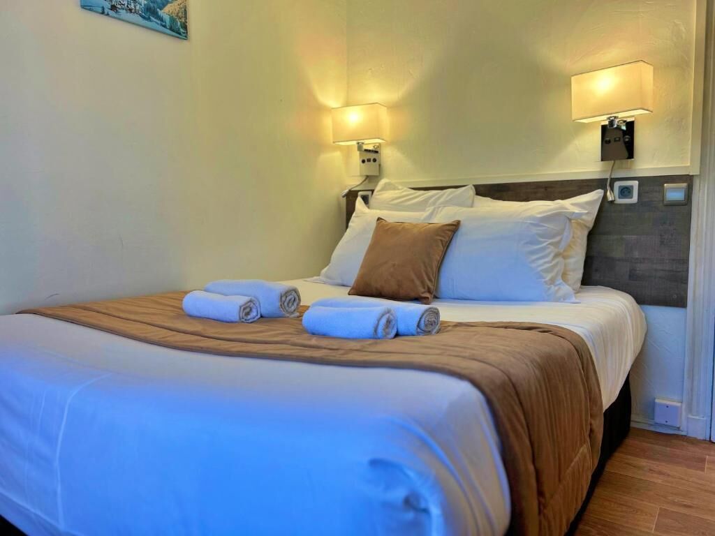 A neatly made bed with white and brown linens, two wall-mounted lamps, and a painting on the wall.
