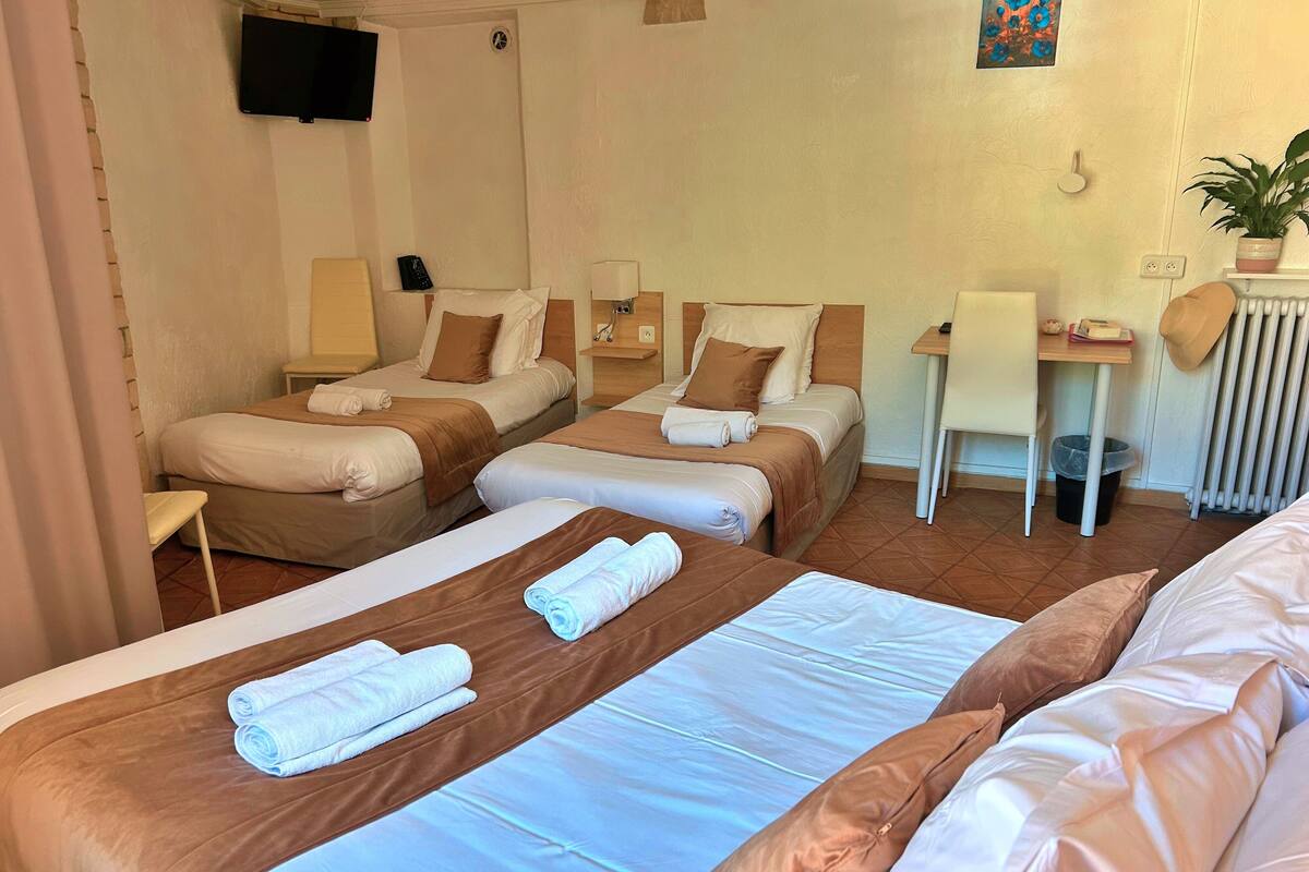 A hotel room with three beds, a desk, a chair, a TV, and a radiator.