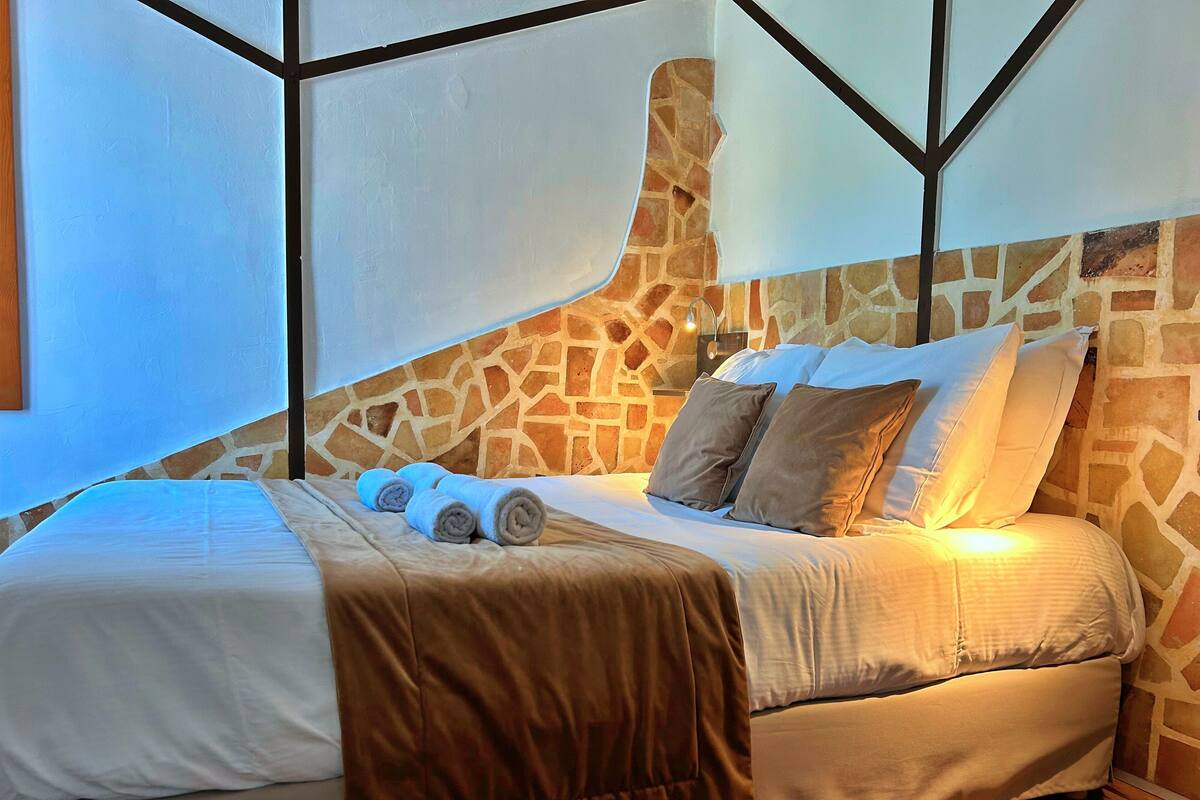 A neatly made bed with rolled towels, a stone wall, and a wooden floor.