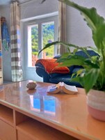Quarto com mesa de madeira, planta em vaso, sofá azul com manta laranja e janela com vista para vegetação.