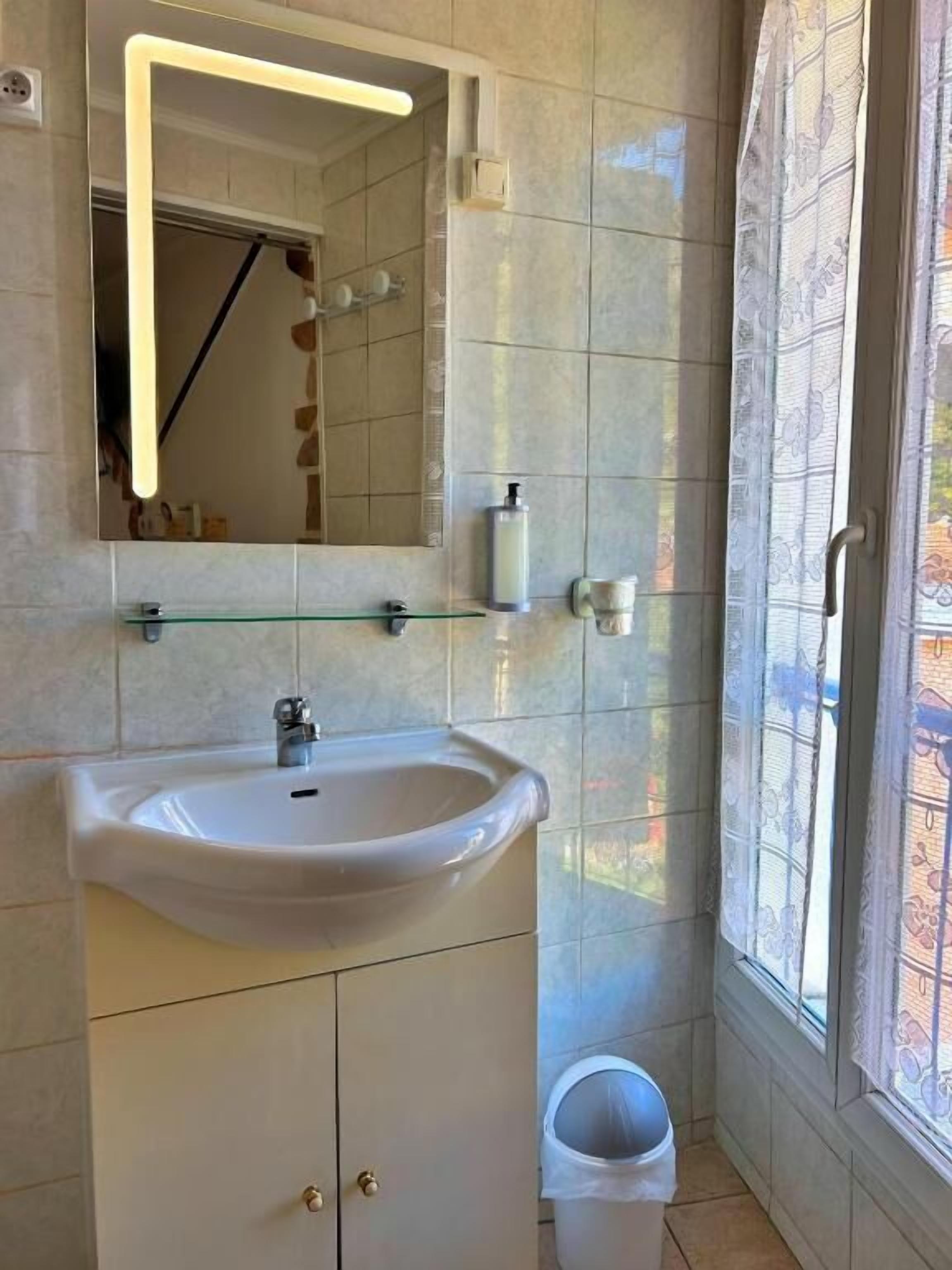 A bathroom with a white sink, mirror, and tiled walls. There is a glass shelf above the sink and a trash bin to the right.