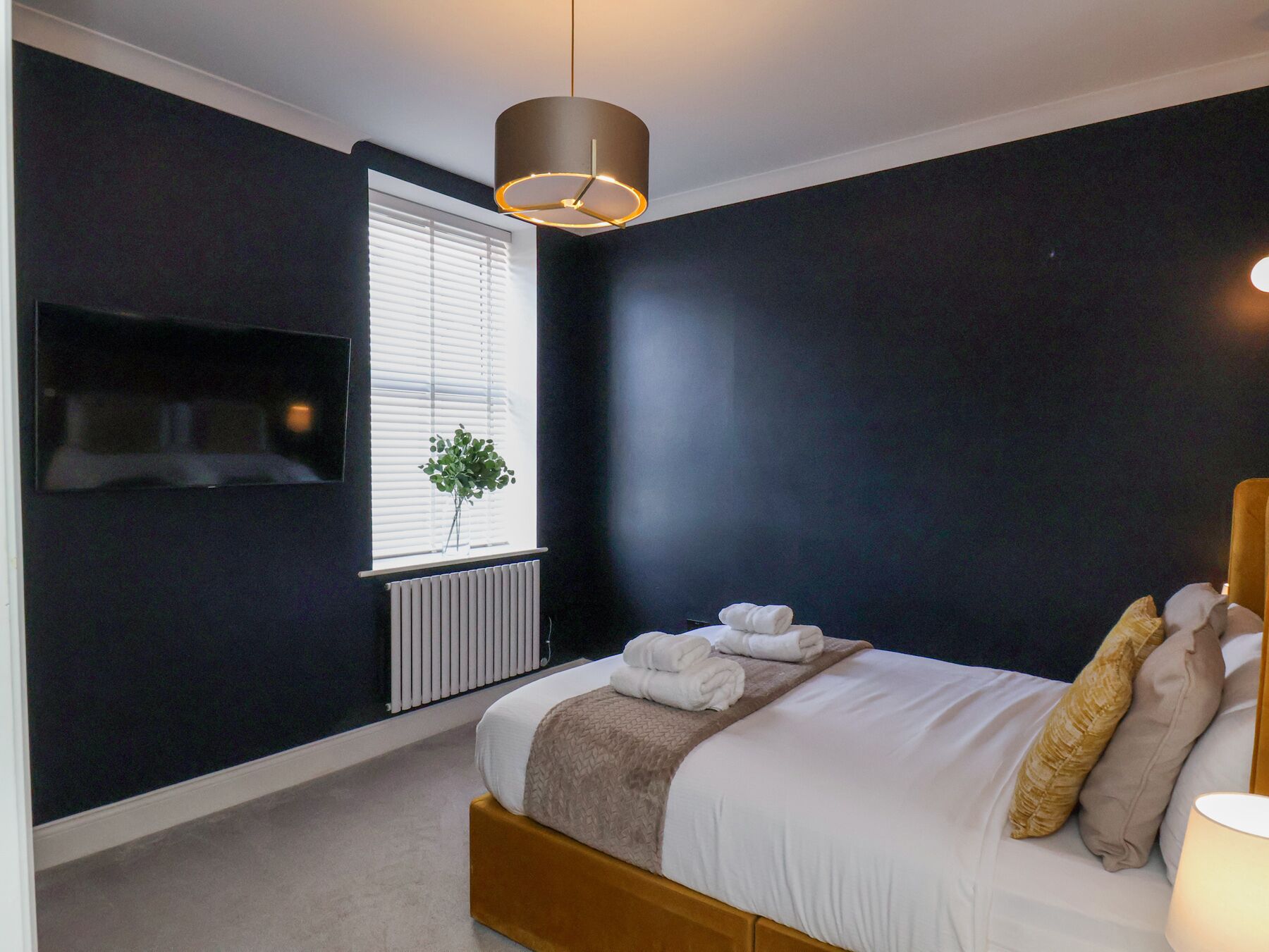 Een moderne slaapkamer met een groot bed, een nachtkastje, een tv aan de muur, een radiator en een raam met jaloezieën.