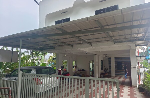 A covered parking area with a car and a motorcycle, a white building with a flat roof, and a tiled floor. - NIRUPAMA HOTEL ALUVA (Aluva)