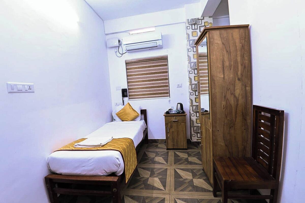 A small, compact room with a bed, a wooden wardrobe, a bedside table, and a wall-mounted air conditioner.