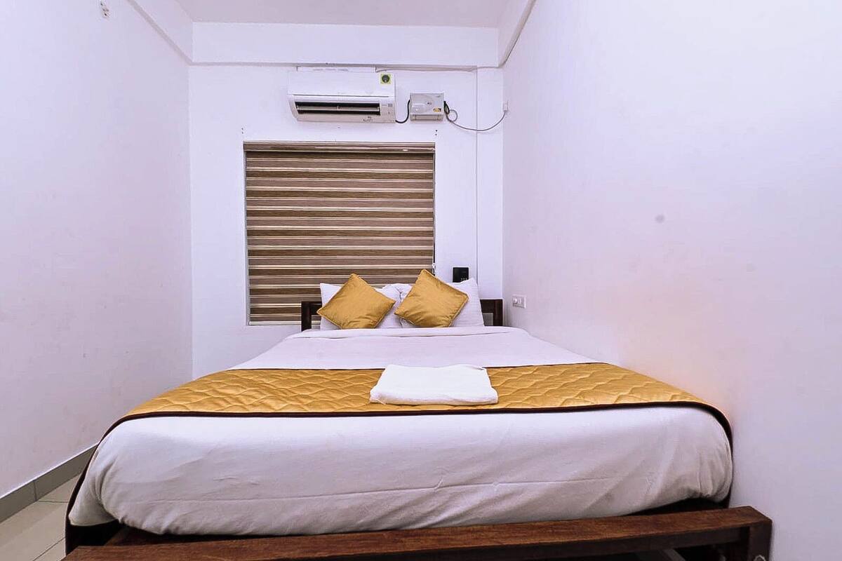 A small, single-bed room with a wooden headboard, a window with blinds, and an air conditioning unit mounted on the wall.